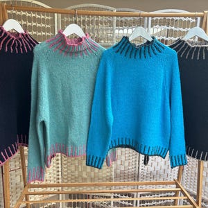 May include: Four long-sleeved knit jumpers hanging on white hangers. The first is black with pink trim, the second is light green with pink trim, the third is bright blue with dark trim, and the fourth is black with light grey trim.