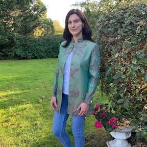 May include: A woman wearing a green jacket with a floral and bird print, blue jeans, and white trainers. She is standing in a grassy area with trees in the background.