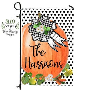 May include: A garden flag with an orange pumpkin design, a black and white checkered bow, and the text "The Harrisons." The background has a black and white polka dot pattern. Green leaves and white flowers are at the bottom.