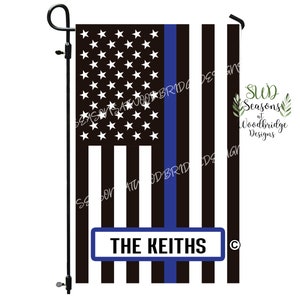 May include: A garden flag featuring a black and white American flag design with a blue stripe. The flag includes the text "THE KEITHS" in a white font on a blue background. The flag is mounted on a black metal stand.