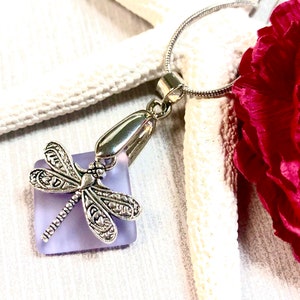 May include: A silver dragonfly charm pendant hangs from a silver chain. The dragonfly is attached to a square piece of lavender glass.