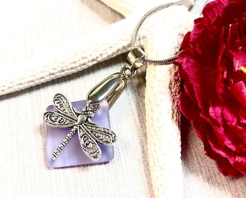 May include: A silver dragonfly charm pendant hangs from a silver chain. The dragonfly is attached to a square piece of lavender glass.
