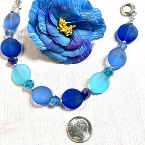 May include: A blue and turquoise glass bead bracelet with silver accents. The bracelet is made with frosted glass beads and crystal accents. The bracelet is shown next to a US dime for size reference.