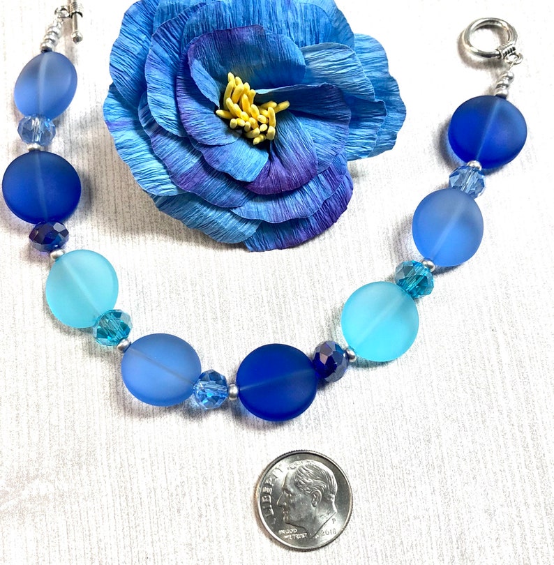 May include: A blue and turquoise glass bead bracelet with silver accents. The bracelet is made with frosted glass beads and crystal accents. The bracelet is shown next to a US dime for size reference.