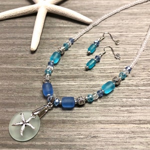May include: A blue and white sea glass necklace with a starfish pendant and matching earrings. The necklace is made with white seed beads and blue sea glass beads.