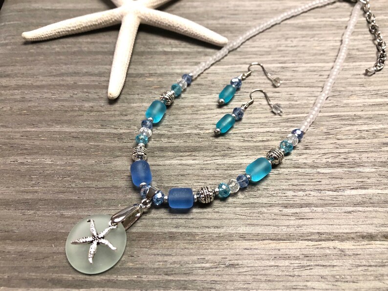 May include: A blue and white sea glass necklace with a starfish pendant and matching earrings. The necklace is made with white seed beads and blue sea glass beads.