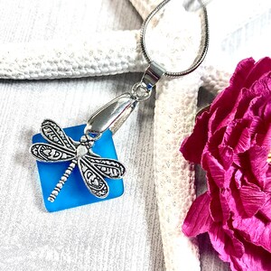 May include: A silver dragonfly charm pendant with a blue glass square. The pendant is on a silver chain and is hanging from a white starfish.