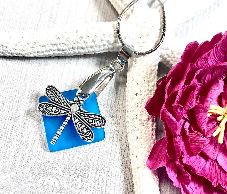 May include: A silver dragonfly charm pendant with a blue glass square. The pendant is on a silver chain and is hanging from a white starfish.