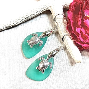 May include: Teal sea glass earrings with silver turtle charms. The earrings are teardrop shaped and dangle from silver hooks.