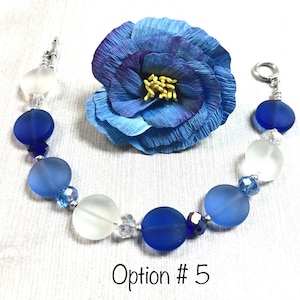 May include: A blue and white glass bead bracelet with a silver clasp. The bracelet features a gradient of blue beads, with the darkest blue beads at the center and the lightest blue beads at the ends. The bracelet also includes clear crystal beads.