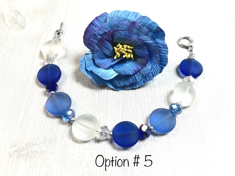 May include: A blue and white glass bead bracelet with a silver clasp. The bracelet features a gradient of blue beads, with the darkest blue beads at the center and the lightest blue beads at the ends. The bracelet also includes clear crystal beads.