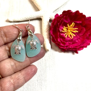 May include: A pair of turquoise sea glass earrings with silver turtle charms. The earrings are dangle style and have a silver hook fastening.