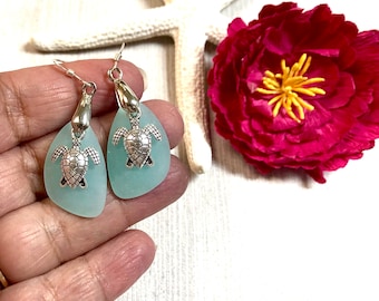 May include: A pair of turquoise sea glass earrings with silver turtle charms. The earrings are dangle style and have a silver hook closure.