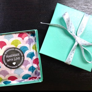 May include: A teal gift box with a white ribbon and white polka dots. The box is open and the inside is lined with colorful patterned tissue paper. A sticker on the tissue paper reads "Handmade with Love".
