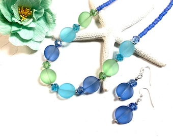 May include: A blue and green glass bead necklace and matching earrings. The necklace has a graduated color scheme, with the beads getting progressively darker from green to blue. The earrings are simple and feature a single blue bead.
