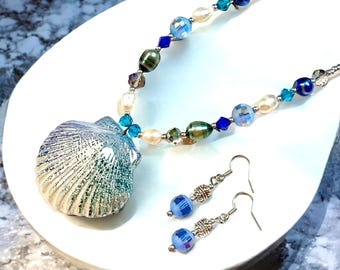 May include: A silver and blue seashell pendant necklace with matching earrings. The necklace features a variety of blue, green, and white beads.