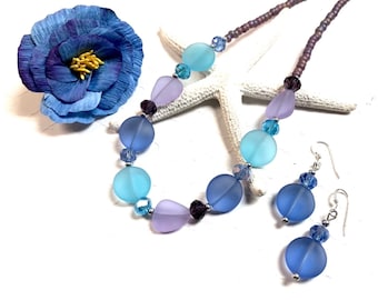 May include: A blue, purple, and light blue glass bead necklace and matching earrings. The necklace is made with frosted glass beads and silver accents. The earrings are dangle earrings with a frosted glass bead and silver accents.