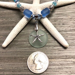 May include: A light blue sea glass necklace with a silver starfish charm. The necklace is made with blue and clear glass beads and silver accents. The charm is a round piece of sea glass with a silver starfish attached to it. The necklace is displayed on a white starfish and a wooden surface.