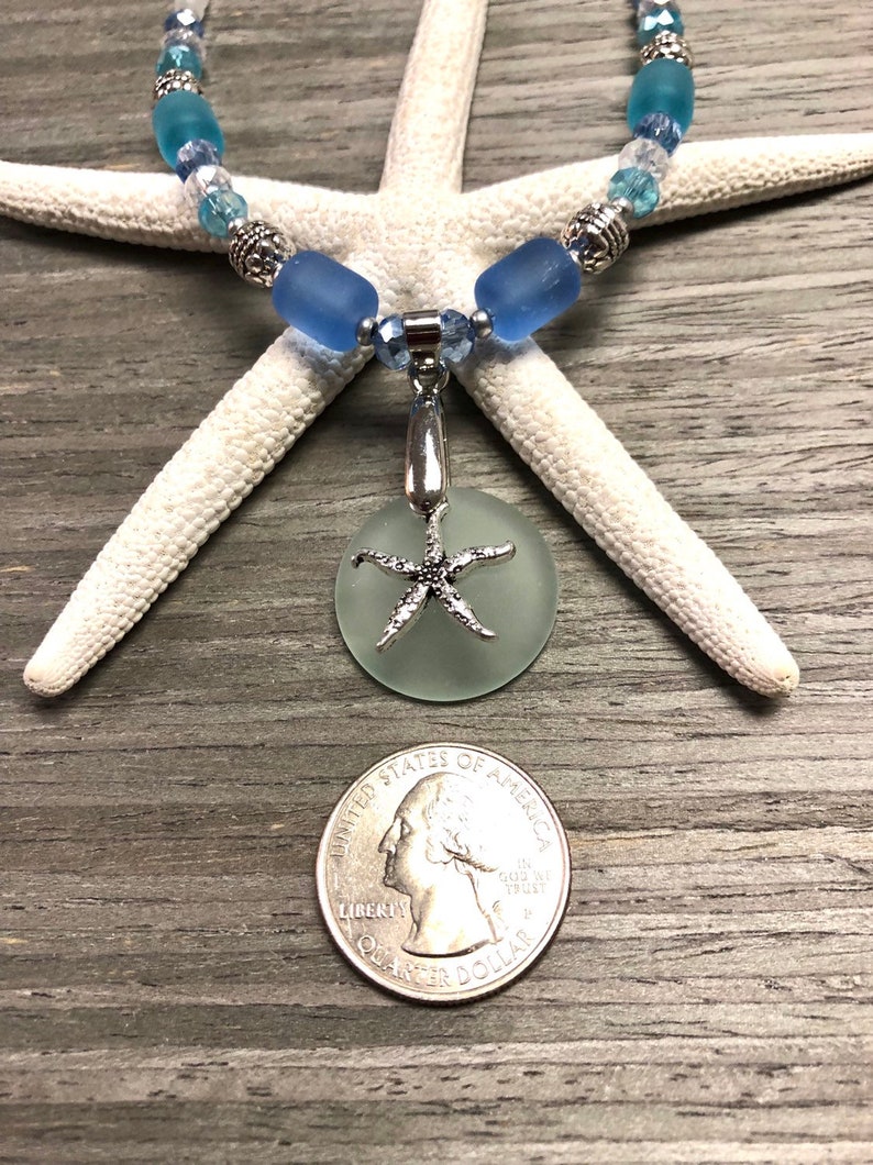 May include: A light blue sea glass necklace with a silver starfish charm. The necklace is made with blue and clear glass beads and silver accents. The charm is a round piece of sea glass with a silver starfish attached to it. The necklace is displayed on a white starfish and a wooden surface.