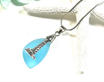 May include: A silver lighthouse charm pendant hangs from a silver chain. The charm is attached to a light blue sea glass teardrop.