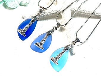May include: Three silver chain necklaces with blue sea glass pendants featuring a silver lighthouse charm. The pendants are teardrop shaped and range in colour from light blue to dark blue.