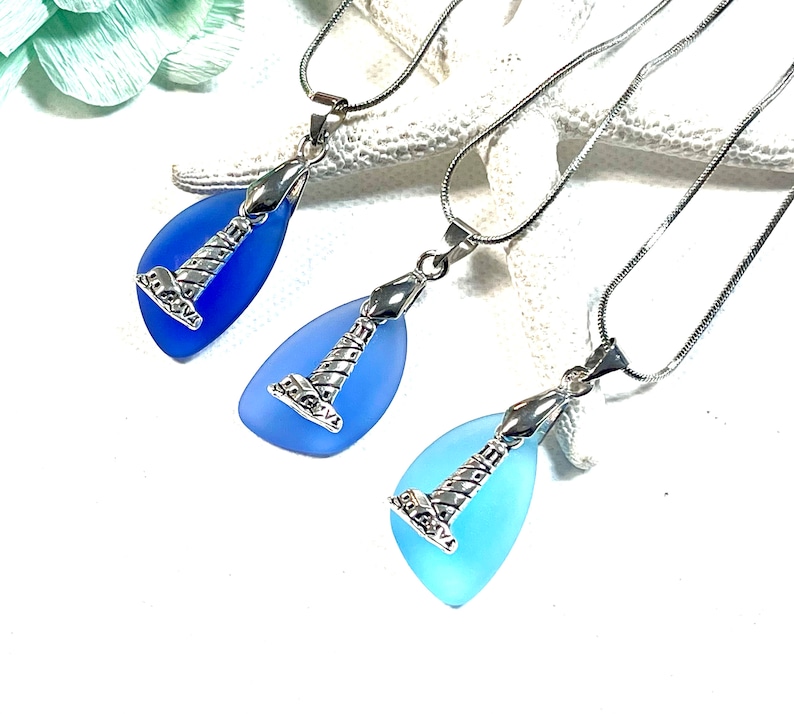 May include: Three silver chain necklaces with blue sea glass pendants featuring a silver lighthouse charm. The pendants are teardrop shaped and range in color from light blue to dark blue.