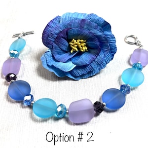May include: A bracelet made with frosted glass beads in shades of blue and purple, separated by small clear crystal beads. The bracelet has a silver toggle clasp.