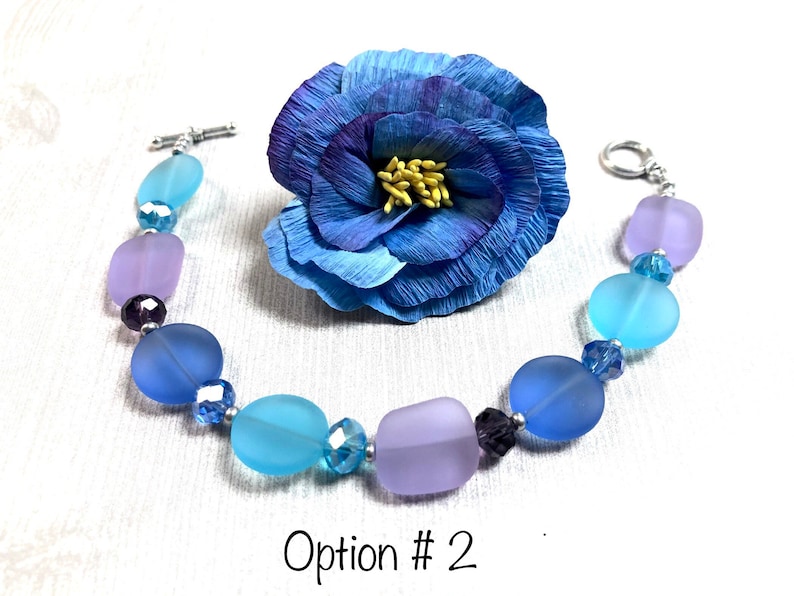 May include: A bracelet made with frosted glass beads in shades of blue and purple, separated by small clear crystal beads. The bracelet has a silver toggle clasp.