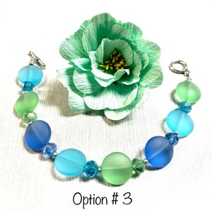 May include: A bracelet with round, frosted glass beads in shades of blue and green. The beads are separated by small silver beads. The bracelet is on a white background with a green paper flower.