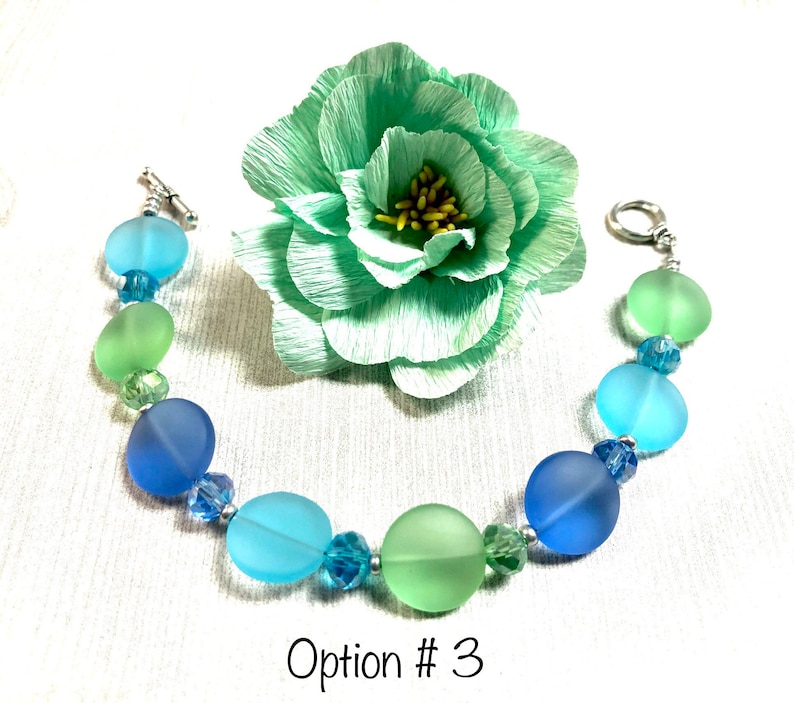 May include: A bracelet with round, frosted glass beads in shades of blue and green. The beads are separated by small silver beads. The bracelet is on a white background with a green paper flower.