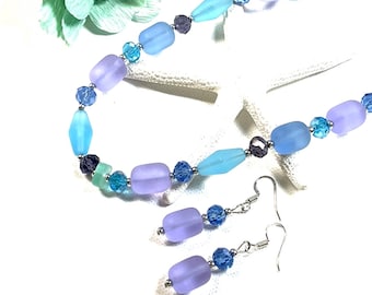May include: A light blue and purple beaded necklace and matching earrings. The necklace features frosted glass beads in shades of blue and purple, with smaller crystal beads interspersed throughout. The earrings are a simple drop style with a single frosted glass bead and a crystal bead.