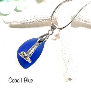 May include: A cobalt blue sea glass pendant necklace with a silver lighthouse charm. The necklace is on a silver chain.