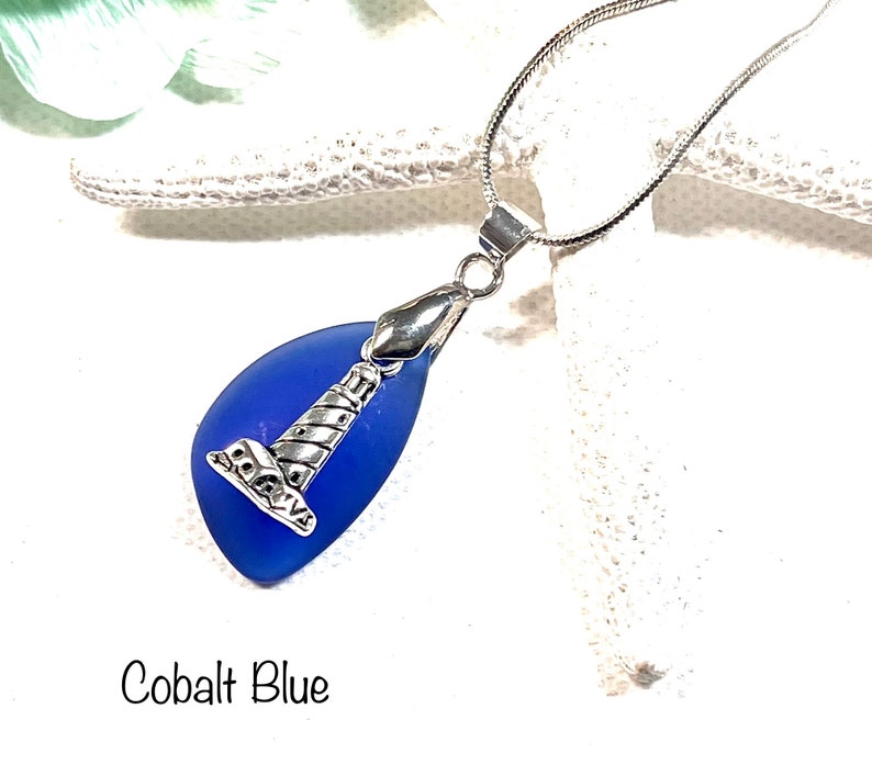 May include: A cobalt blue sea glass pendant necklace with a silver lighthouse charm. The necklace is on a silver chain.