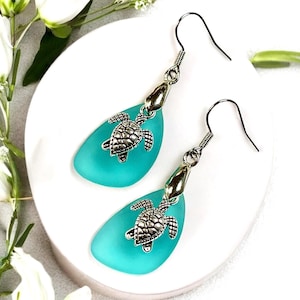 May include: Teal blue glass teardrop earrings with silver turtle charms. The earrings have silver hooks.