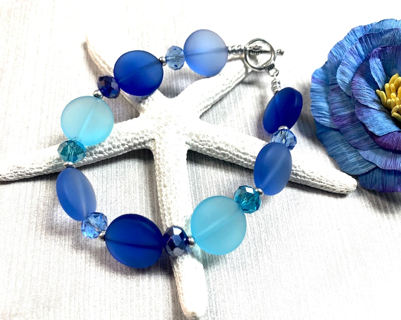 May include: A blue and light blue glass bead bracelet with a silver clasp. The bracelet is made with frosted glass beads and crystal accents.