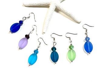 May include: Five pairs of dangle earrings with oval-shaped glass beads in various shades of blue, green, and purple. Each earring features a small crystal charm.