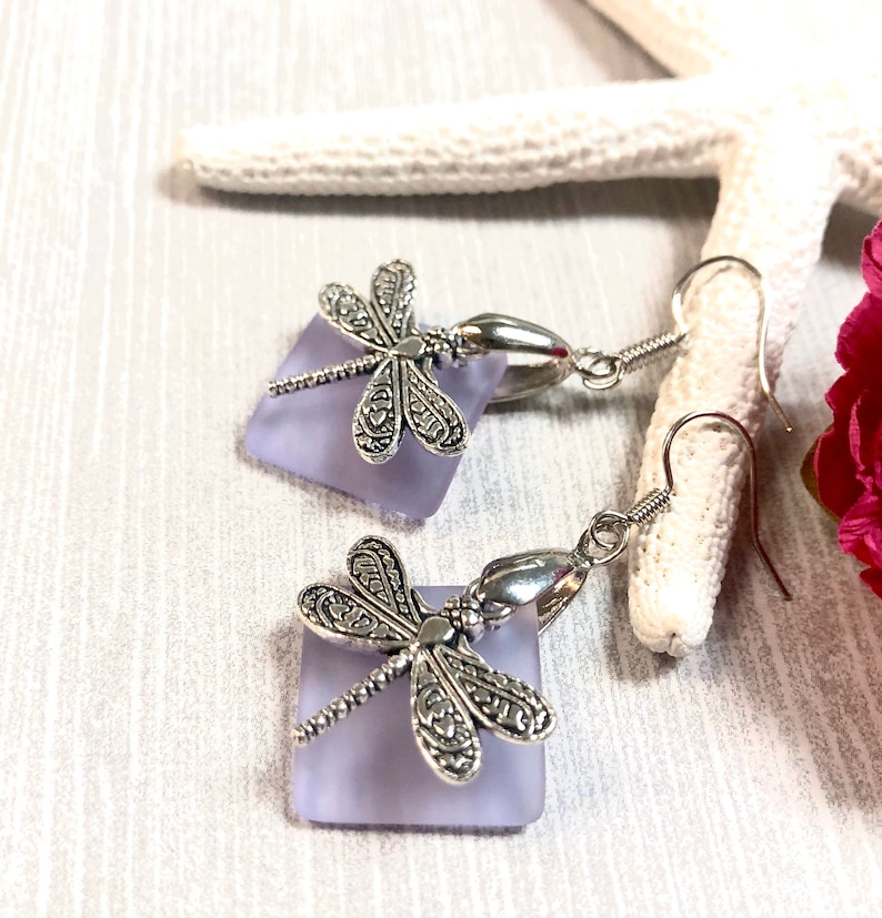 May include: A pair of silver dragonfly earrings with lavender glass squares. The earrings have a hook closure.