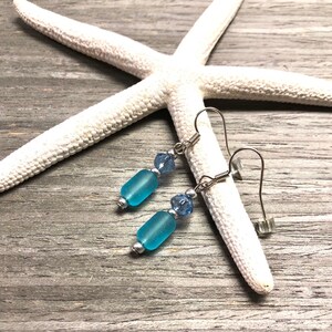May include: A pair of dangle earrings with light blue glass beads and silver accents. The earrings are on a white starfish on a wood background.