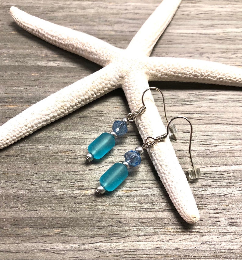May include: A pair of dangle earrings with light blue glass beads and silver accents. The earrings are on a white starfish on a wood background.