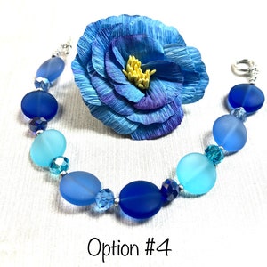 May include: A blue and light blue glass bead bracelet with a silver clasp. The bracelet has a gradient of blue colors, with the darkest blue beads at the top and the lightest blue beads at the bottom. The bracelet is accented with clear crystal beads.