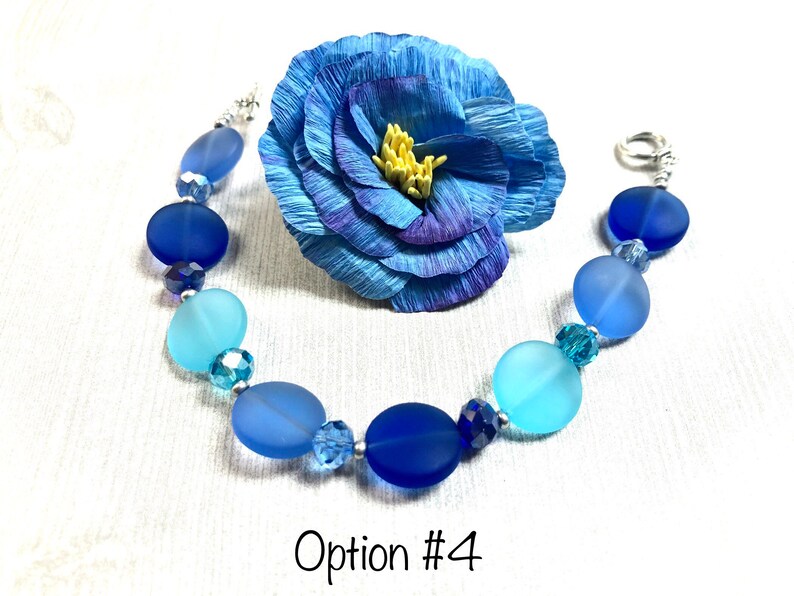 May include: A blue and light blue glass bead bracelet with a silver clasp. The bracelet has a gradient of blue colors, with the darkest blue beads at the top and the lightest blue beads at the bottom. The bracelet is accented with clear crystal beads.