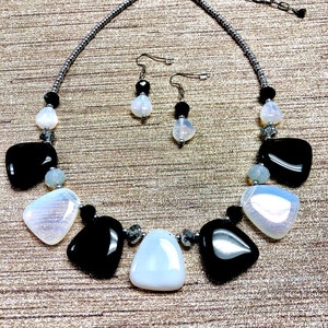 May include: A black and white statement necklace and matching earrings set. The necklace features large, irregular shaped black and white stones with silver accents. The earrings are simple and feature a single black stone with a white stone accent.