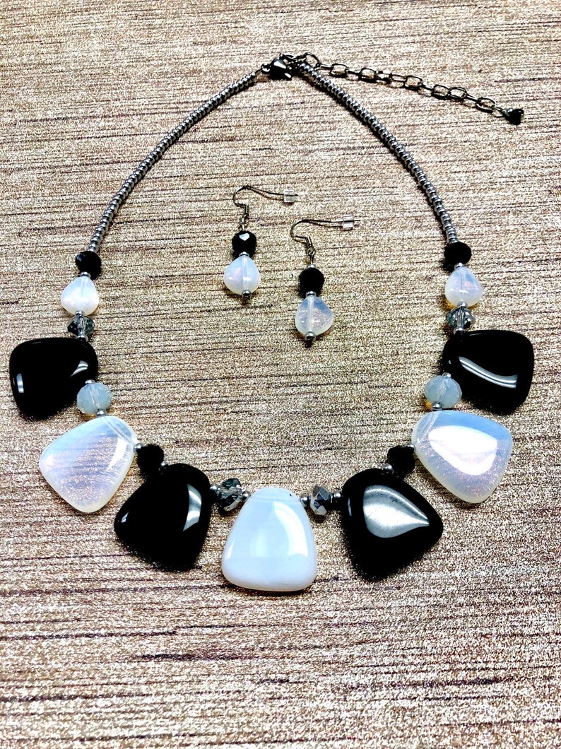 May include: A black and white statement necklace and matching earrings set. The necklace features large, irregular shaped black and white stones with silver accents. The earrings are simple and feature a single black stone with a white stone accent.