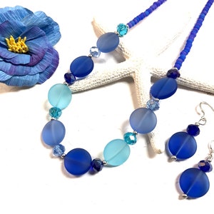 May include: A blue and turquoise glass bead necklace and matching earrings. The necklace features a graduated ombre effect with larger, round beads. The earrings are simple and feature a single round bead.