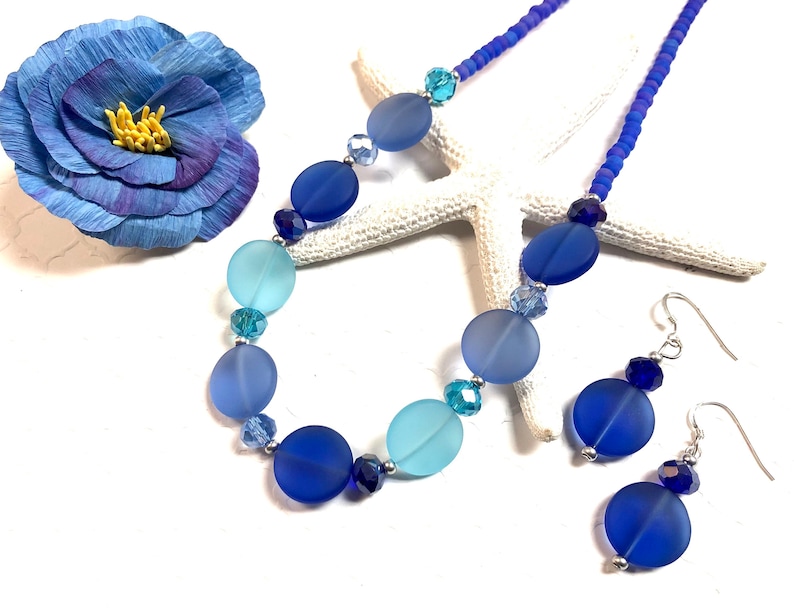 May include: A blue and turquoise glass bead necklace and matching earrings. The necklace features a graduated ombre effect with larger, round beads. The earrings are simple and feature a single round bead.