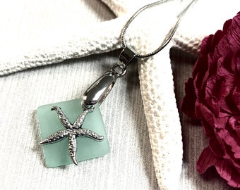 May include: A silver starfish charm pendant necklace with a light blue square glass charm. The necklace is on a white starfish and a red flower.