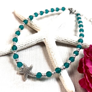 May include: Teal blue glass bead anklet with a silver starfish charm. The anklet is on a white background with a starfish and a pink flower.