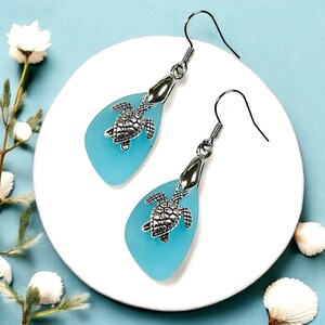 Peut inclure: Une paire de boucles d'oreilles pendantes avec un charme de tortue argentée et une pièce de verre de mer bleu clair en forme de larme.