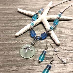 May include: A blue and white sea glass necklace and earrings set with a starfish charm. The necklace features blue and white beads and a round sea glass pendant with a silver starfish charm. The earrings feature blue sea glass beads and silver hooks.