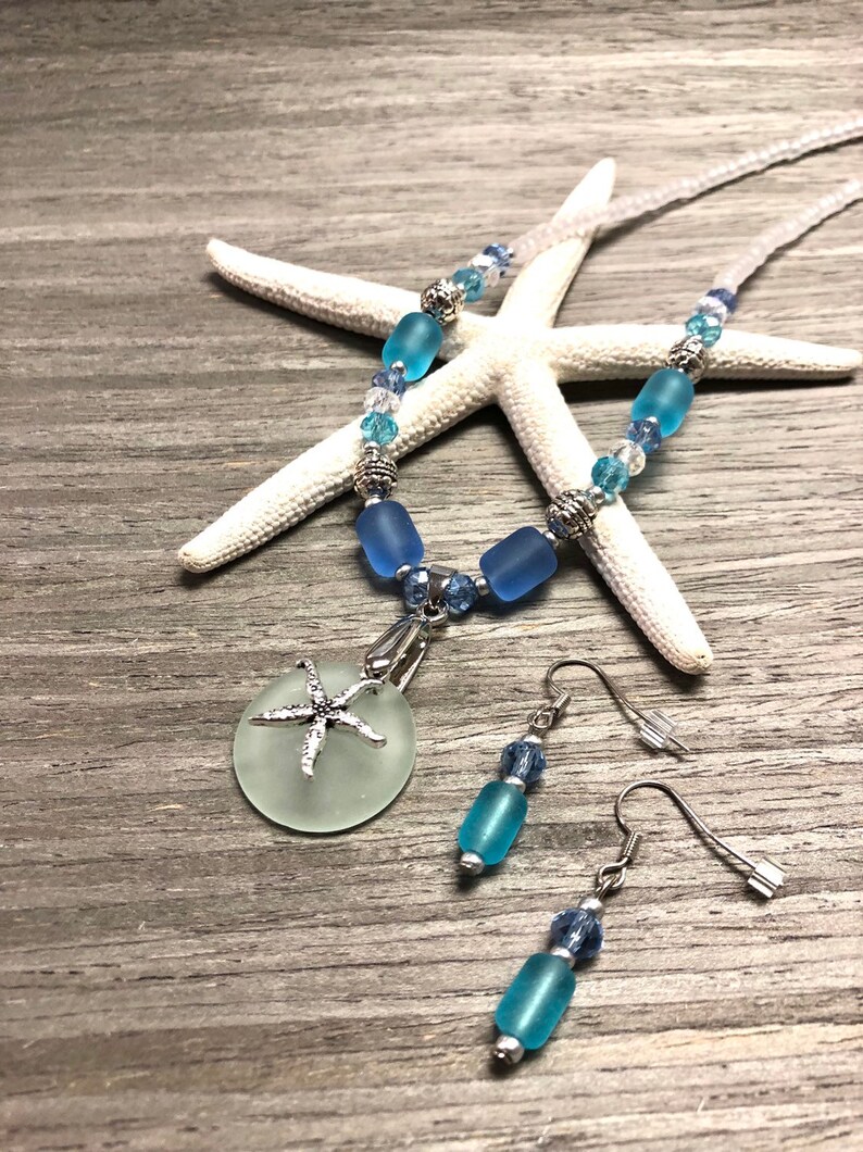 May include: A blue and white sea glass necklace and earrings set with a starfish charm. The necklace features blue and white beads and a round sea glass pendant with a silver starfish charm. The earrings feature blue sea glass beads and silver hooks.